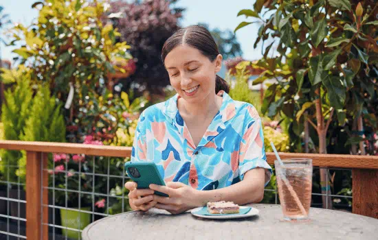 Woman sitting on an outdoor deck while on her phone. On the table in front of her is a plate with a pastry on it and a glass containing a beverage in it. The background shows a variety of plants and trees.