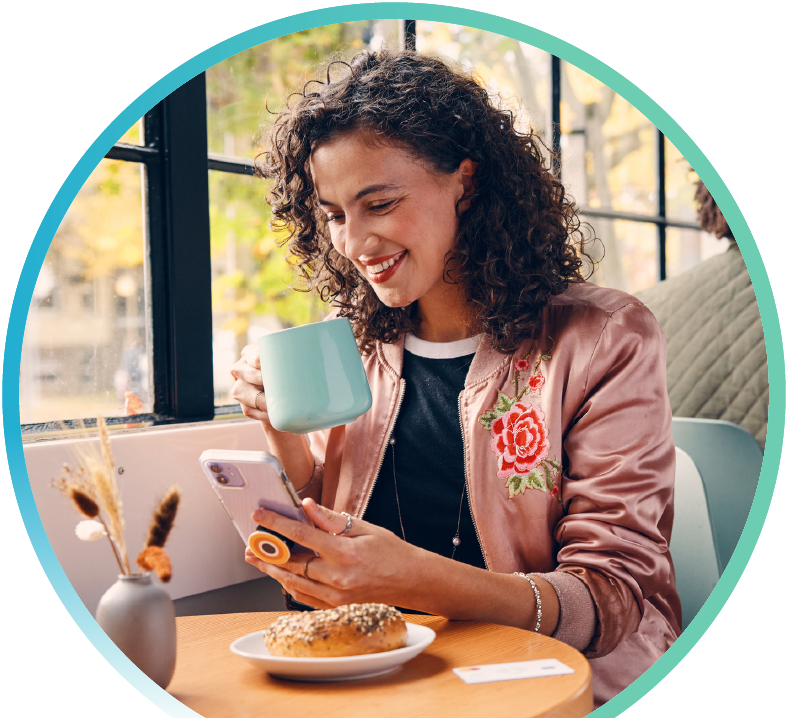 Person drinking out of a mug while browsing there phone and eating a bagel.