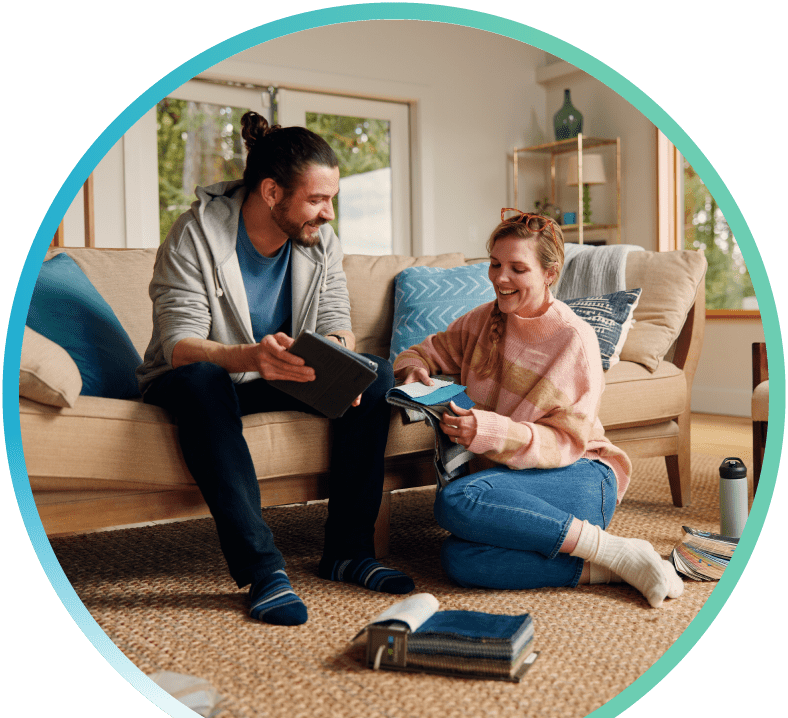 Couple sitting together in a living room setting. The man is sitting on the sofa holding a tablet, while the woman is sitting on the floor beside him holding a booklet.