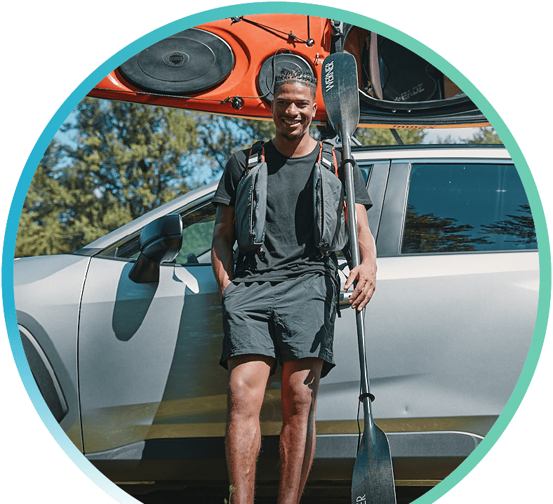 Picture of a man leaning on their car while holding a kayak paddle and a kayak strapped to the top of their car.