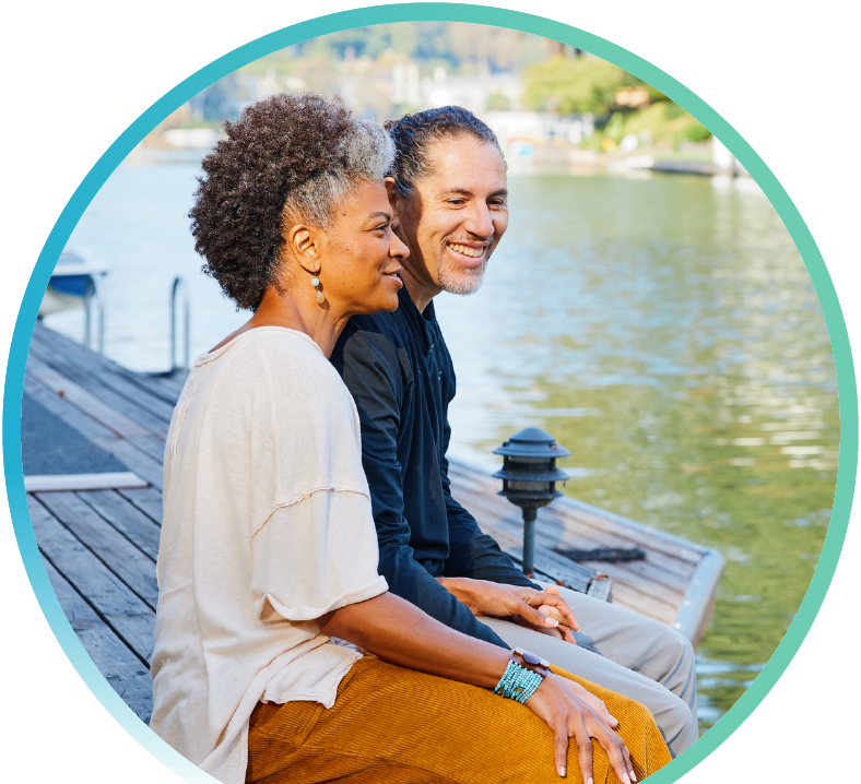 A couple sitting on a deck with their legs dangling off the edge and above a body of water.