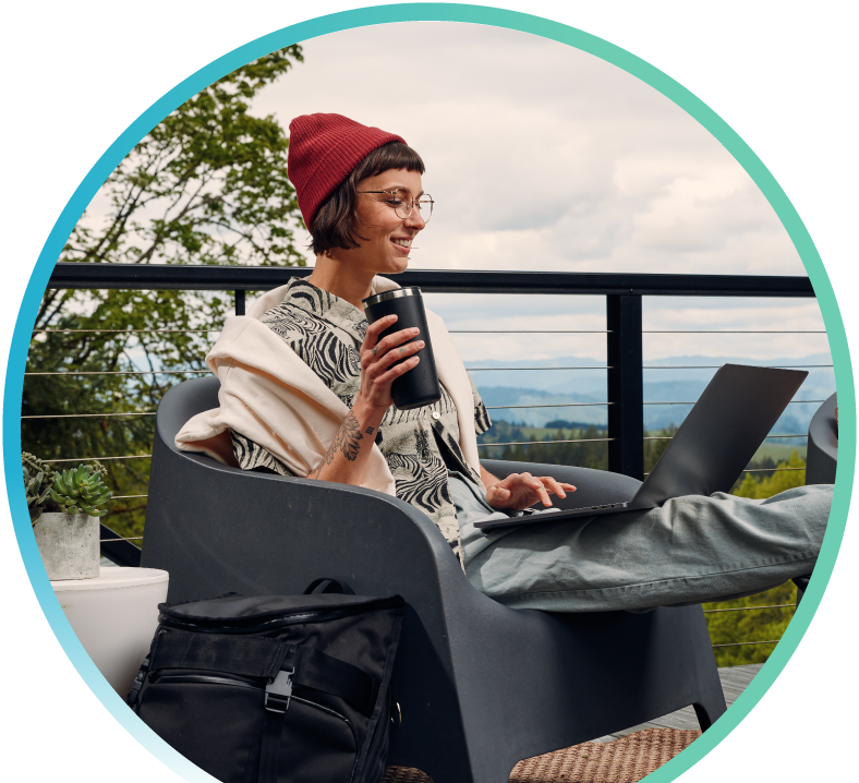 Woman sitting on an outdoor balcony while holding a tumbler in one hand and typing on her laptop with the other hand.