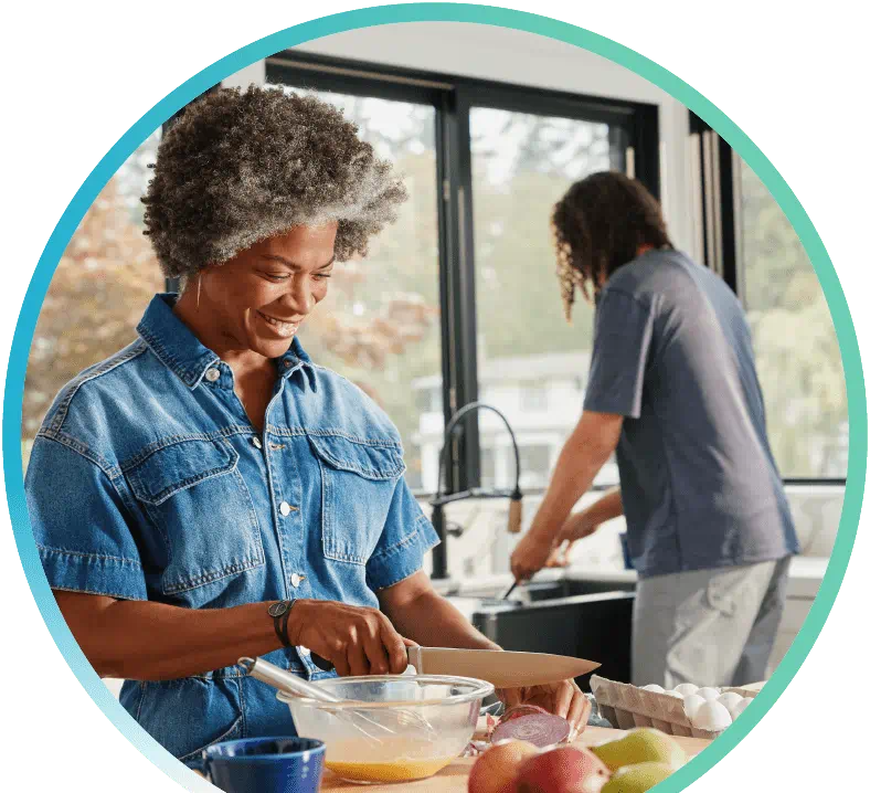 Woman preparing food in a kitchen, while a man is cleaning dishes in the background.