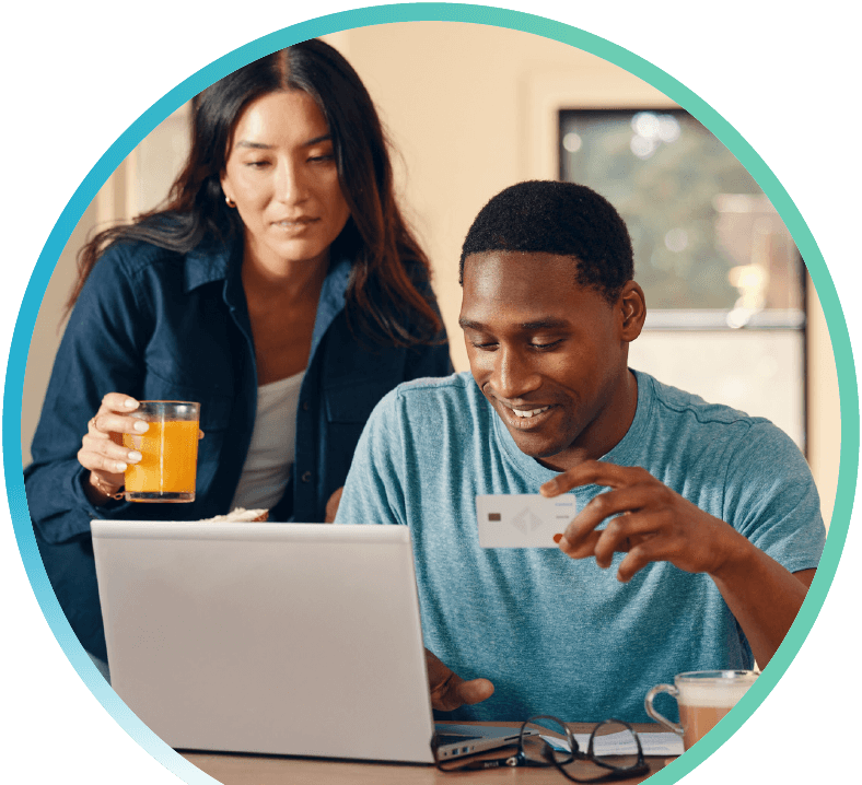 Man and woman looking at a laptop. Woman is standing and holding a drink, the man is sitting and holding a credit card