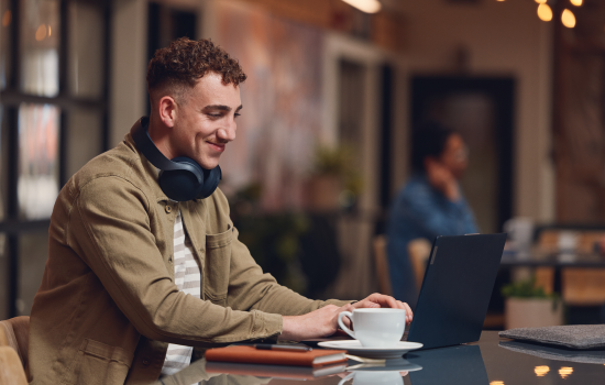 A man sitting in a coffee shop while working on his laptop. Beside him is a coffee mug with headphones wrapped around his neck..