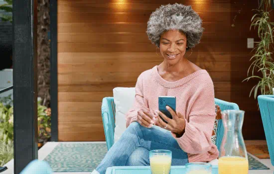 A woman sitting in a sun room while on her phone. On the table in front of her are a few glasses with what appears to be lemonade or some other type of beverage.